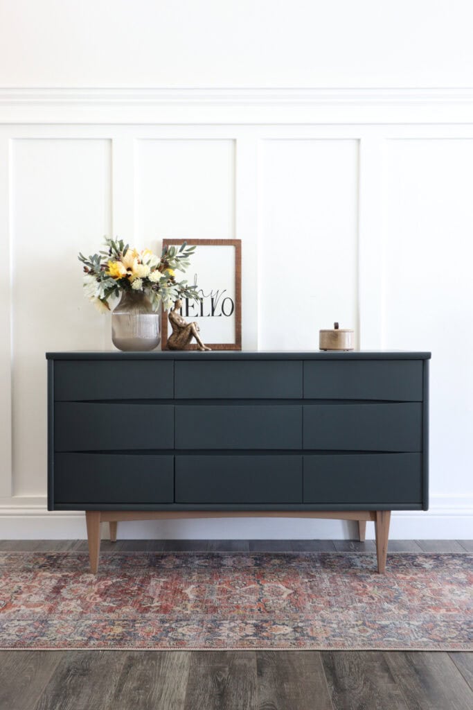 Dark green dresser with wooden legs, a vase of flowers, framed hello sign, and decor on top.