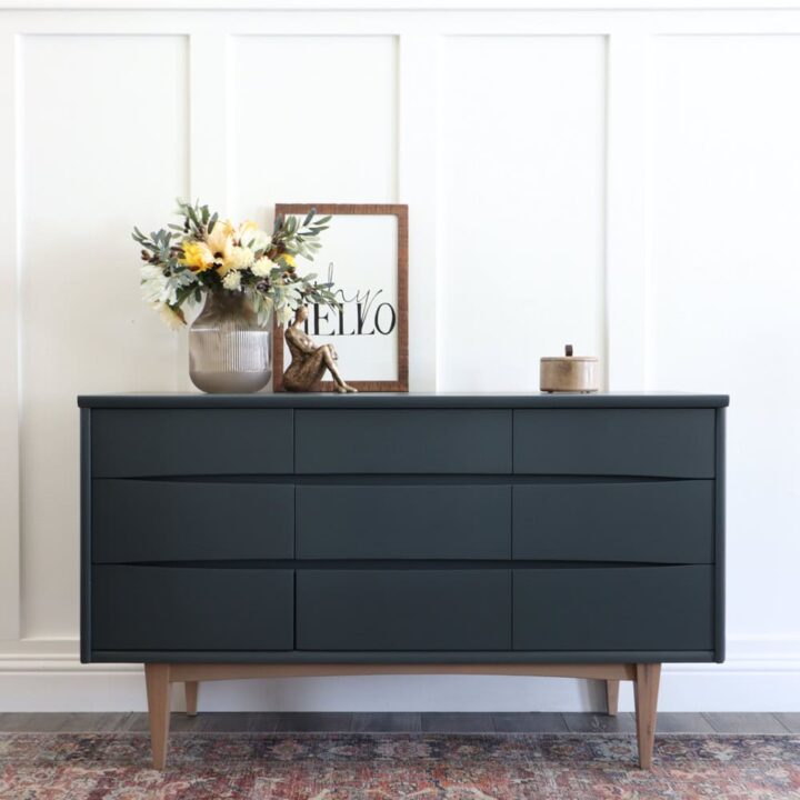 Dark green dresser with wooden legs, a vase of flowers, framed hello sign, and decor on top.