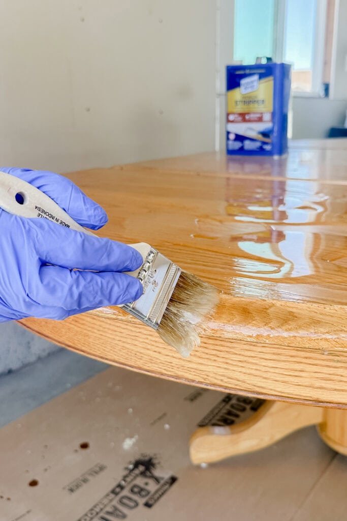 Using a paintbrush to apply chemical stripper to a wooden table surface.