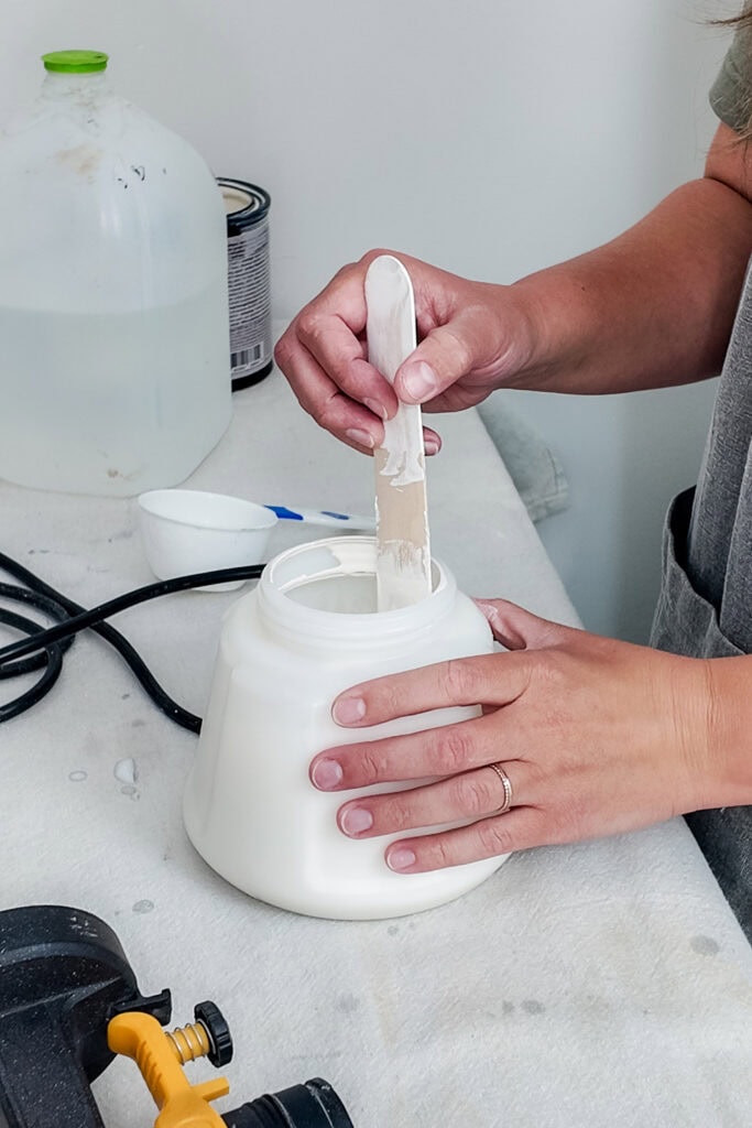 A person stirs white paint in sprayer container with a wooden stick