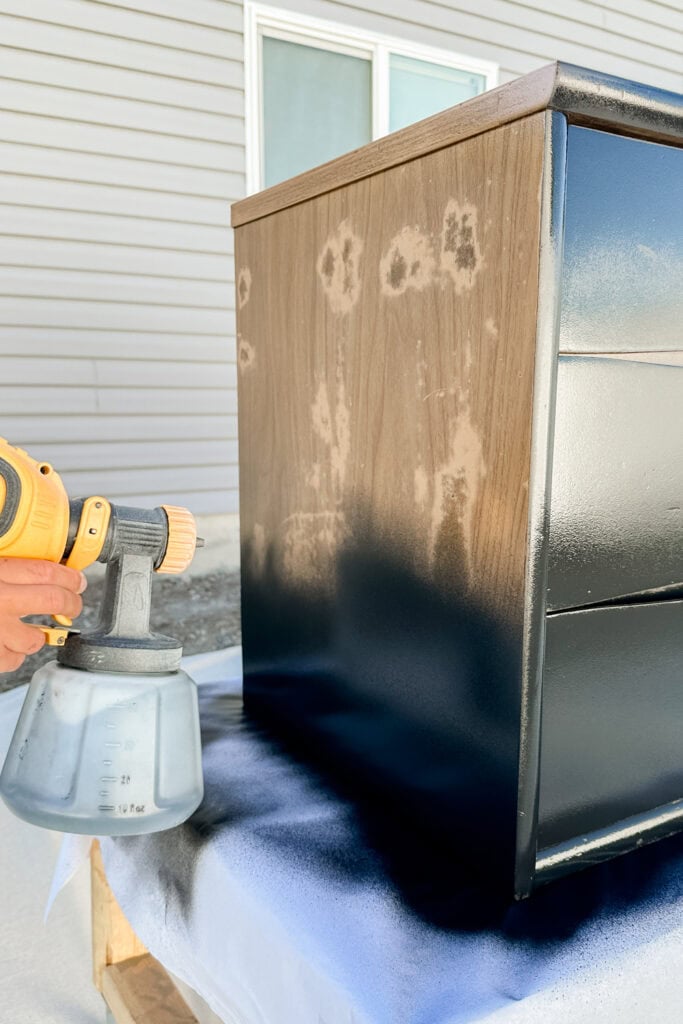 A hand uses a paint sprayer to apply black primer to a wooden dresser outdoors