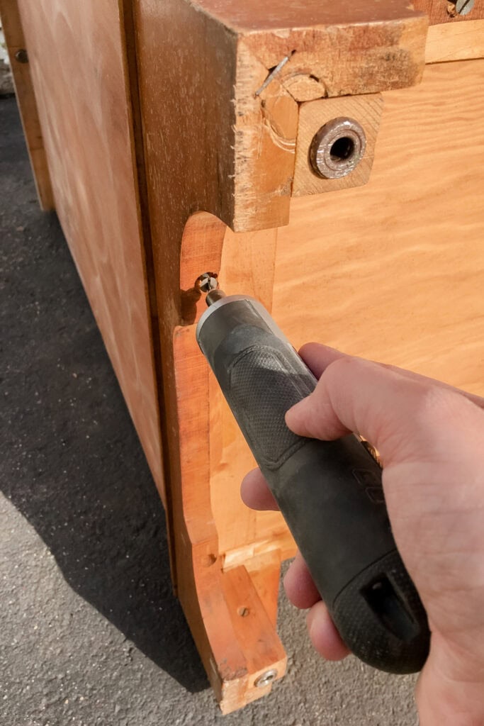 Hand using a black electric screwdriver to remove a screw from a wooden furniture base.