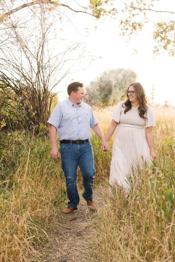 A couple walks hand in hand through a grassy field, smiling at each other on a sunny day.