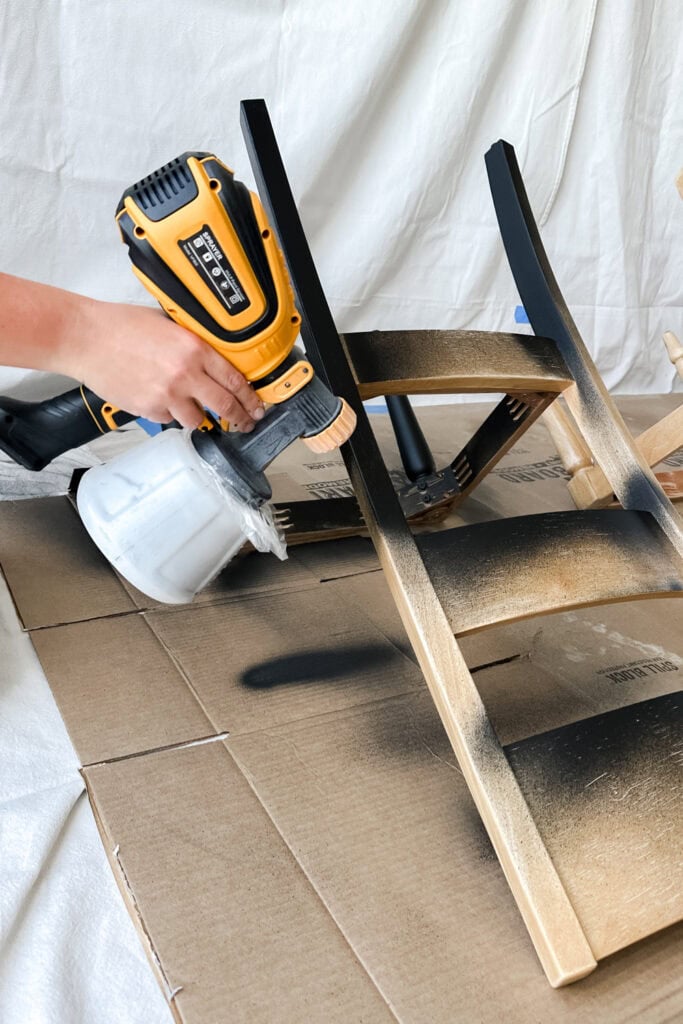 A hand uses a paint sprayer to apply black primer on chair