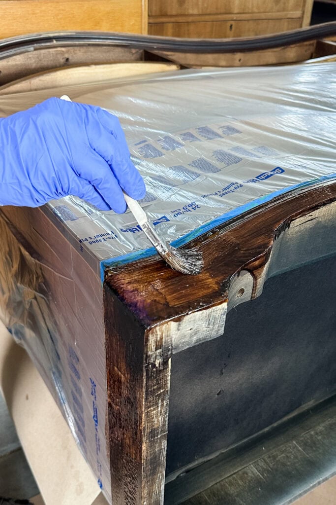 A gloved hand applies wood stain to a furniture edge with a brush