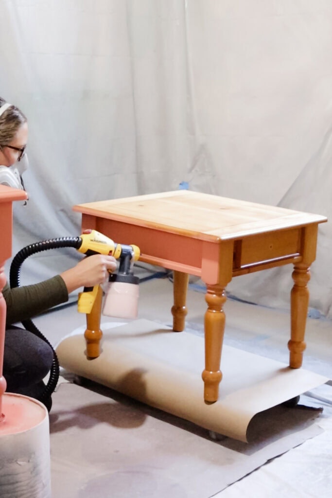 Person wearing respirator applying coral paint onto table with sprayer