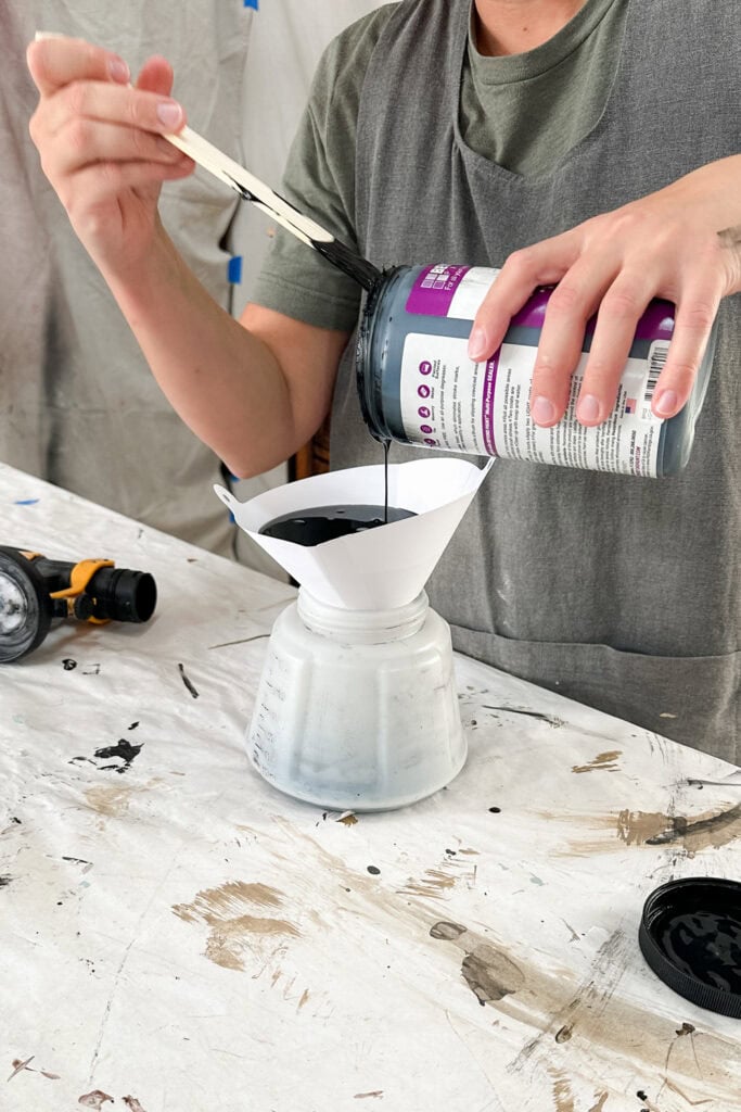 A person pours black paint through a filter into a sprayer container