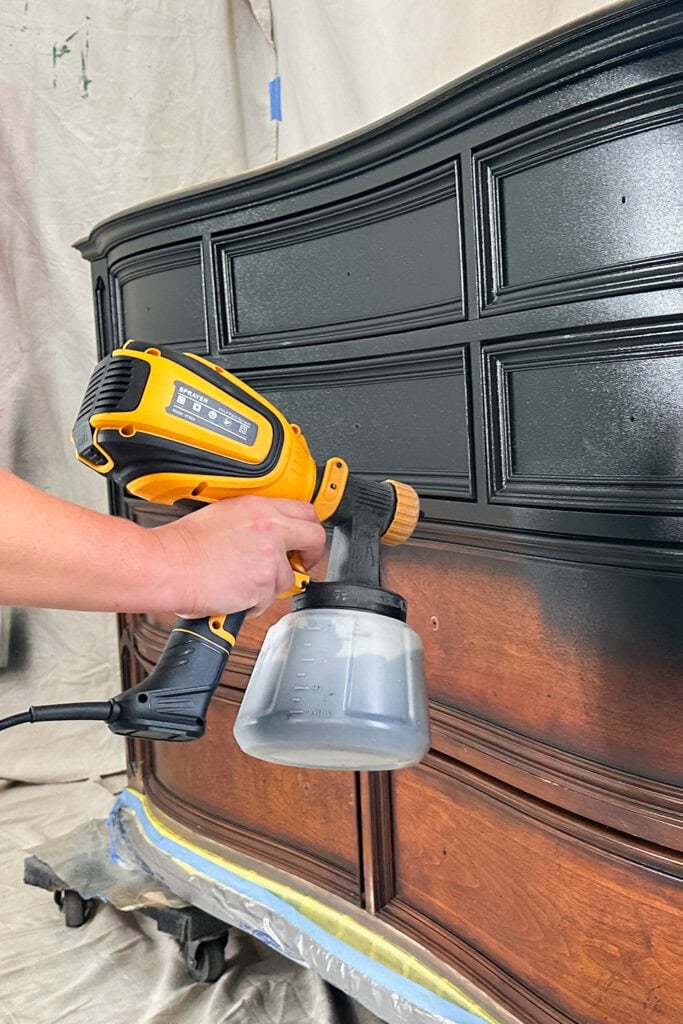 A person spray paints a wooden dresser black with a handheld sprayer.