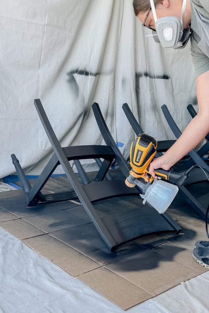 A person wearing a respirator spray paints black chairs in a protected workspace.