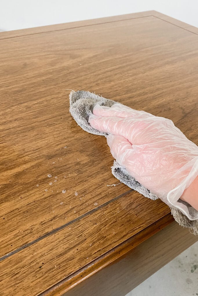 A hand in a plastic glove wipes a wooden table with a gray cloth.