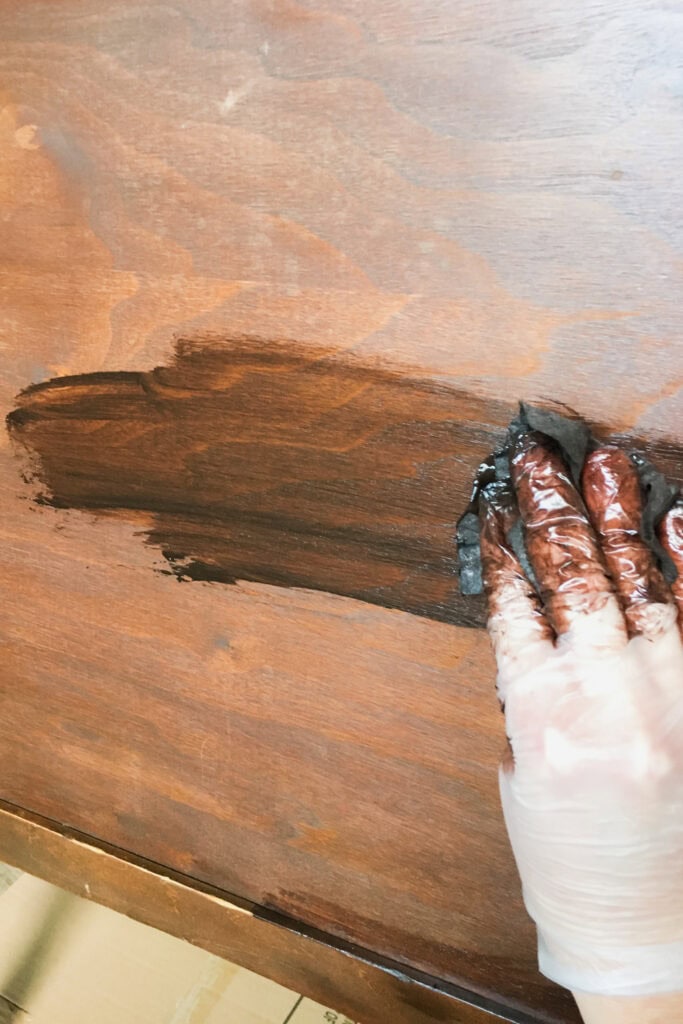 A gloved hand applies dark gel stain to a wooden surface using a cloth.