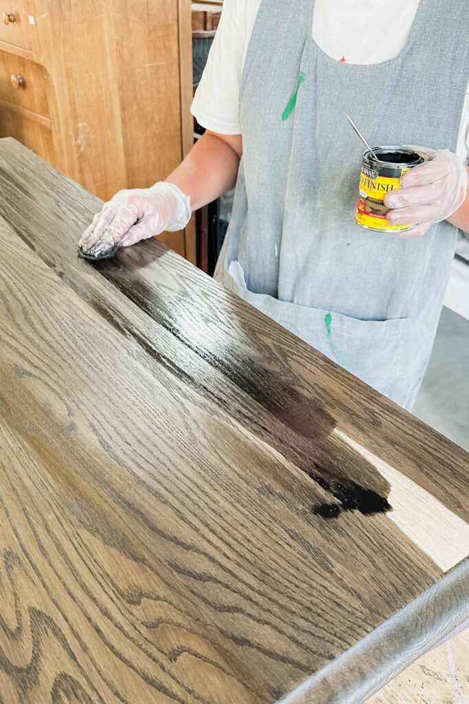 Person staining a wooden surface with dark wood stain, wearing gloves and an apron.