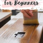 A hand holds a brush to apply stain on a wooden furniture piece, with text: How to Restain Furniture for Beginners.