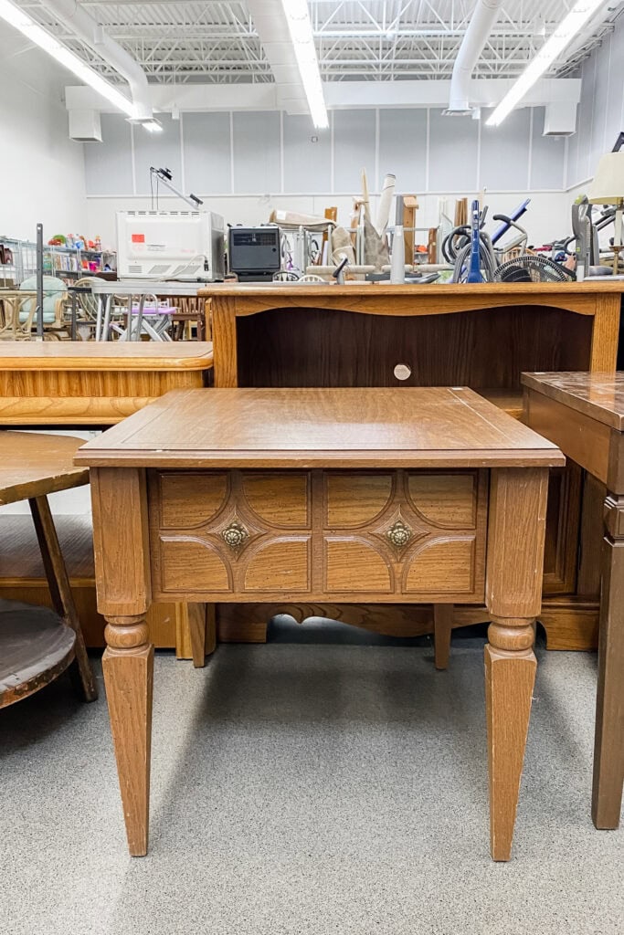 A square wooden table with carved details and brass knobs, displayed in a thrift store.