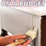 Hand painting white paint on a wooden piece of furniture with text How to Paint Furniture on a Budget.