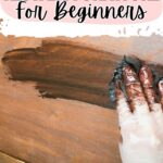 Gloved hand applying stain to wood with the text “How to Restain Furniture For Beginners” at the top.
