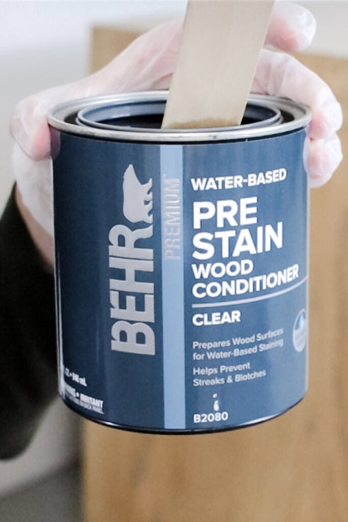 A hand holding a can of BEHR Pre Stain Wood Conditioner with a wooden stick inside.