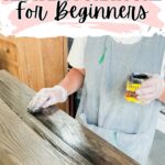 Person wearing gloves and apron applies dark wood stain to a tabletop with a cloth. Text: How to Restain Furniture for Beginners.