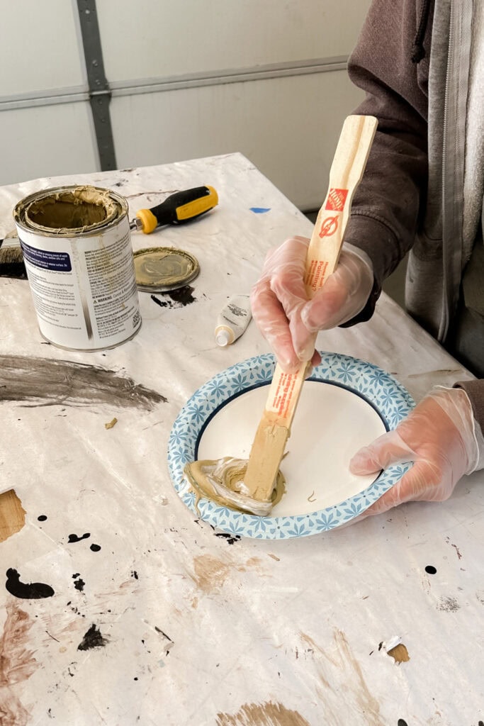 Gloved hands mix Bondo putty on a paper plate with a wooden stick