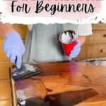 Person in gloves applies dark stain to wooden furniture with a scraper; text reads How to Restain Furniture For Beginners.