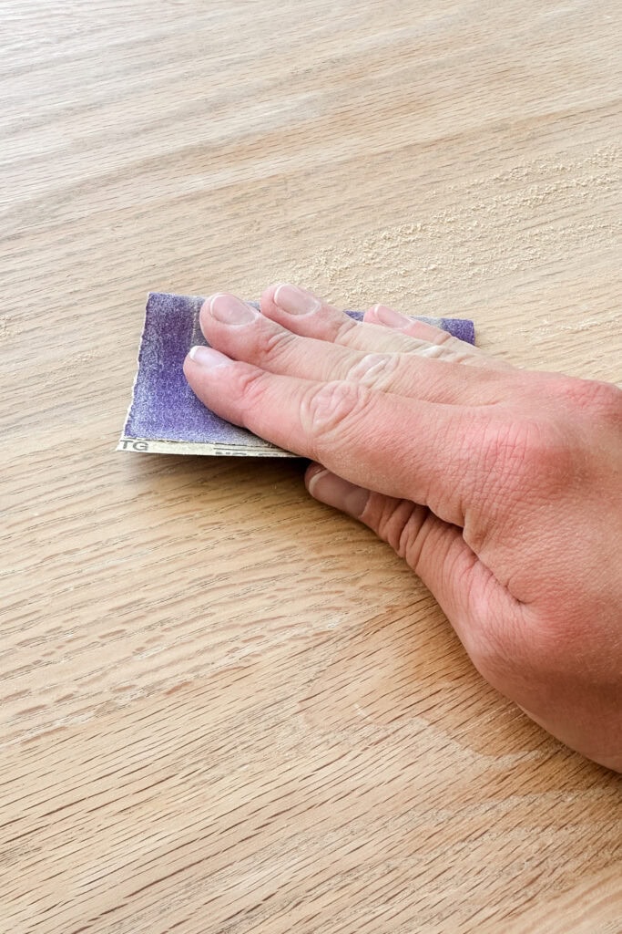 A hand sanding a wooden surface with a piece of sandpaper.