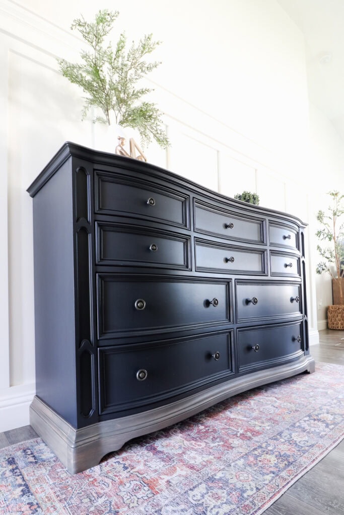 A black dresser with nine drawers stands on a rug with plants and decor on top