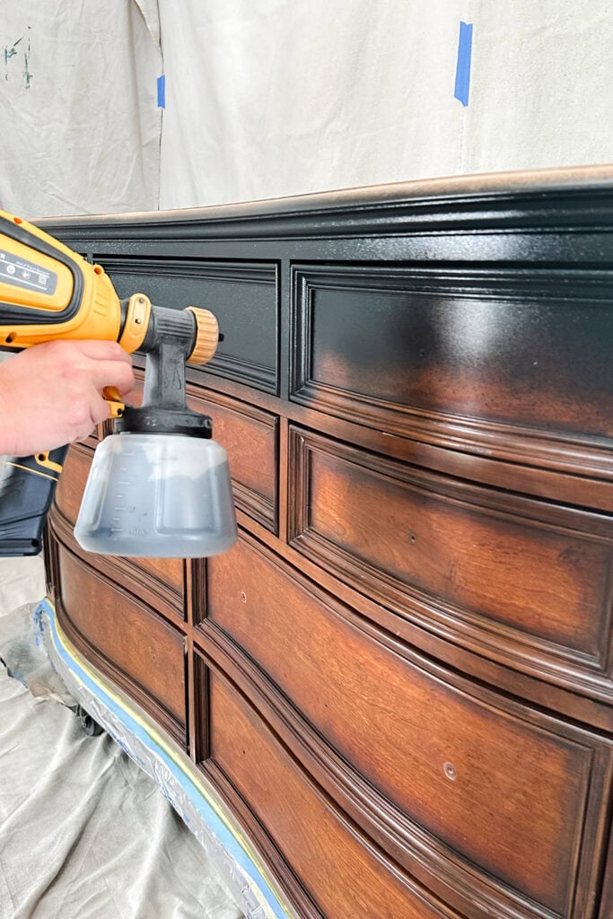 A person spray paints the top drawers of a wooden dresser black