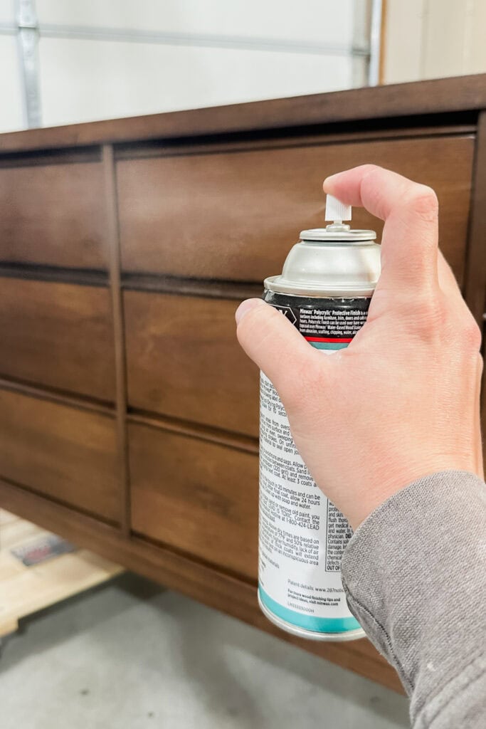A hand sprays a can of topcoat onto a wooden dresser