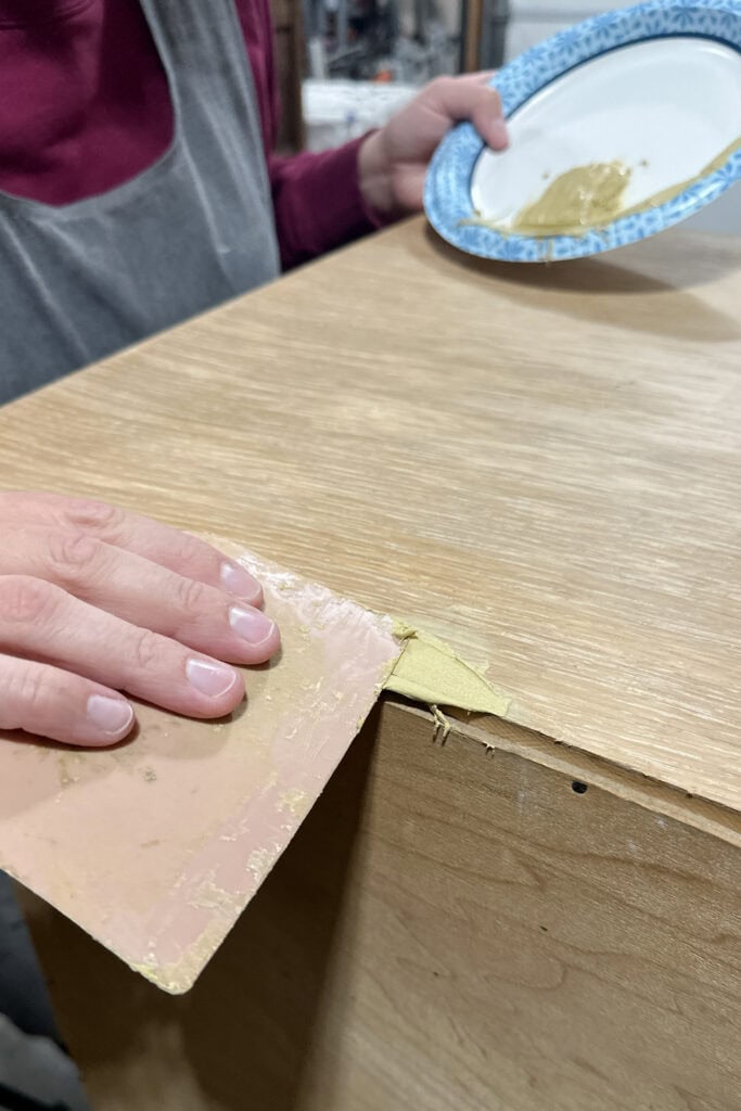 A person applies wood filler to a corner of a wooden surface using a putty knife.