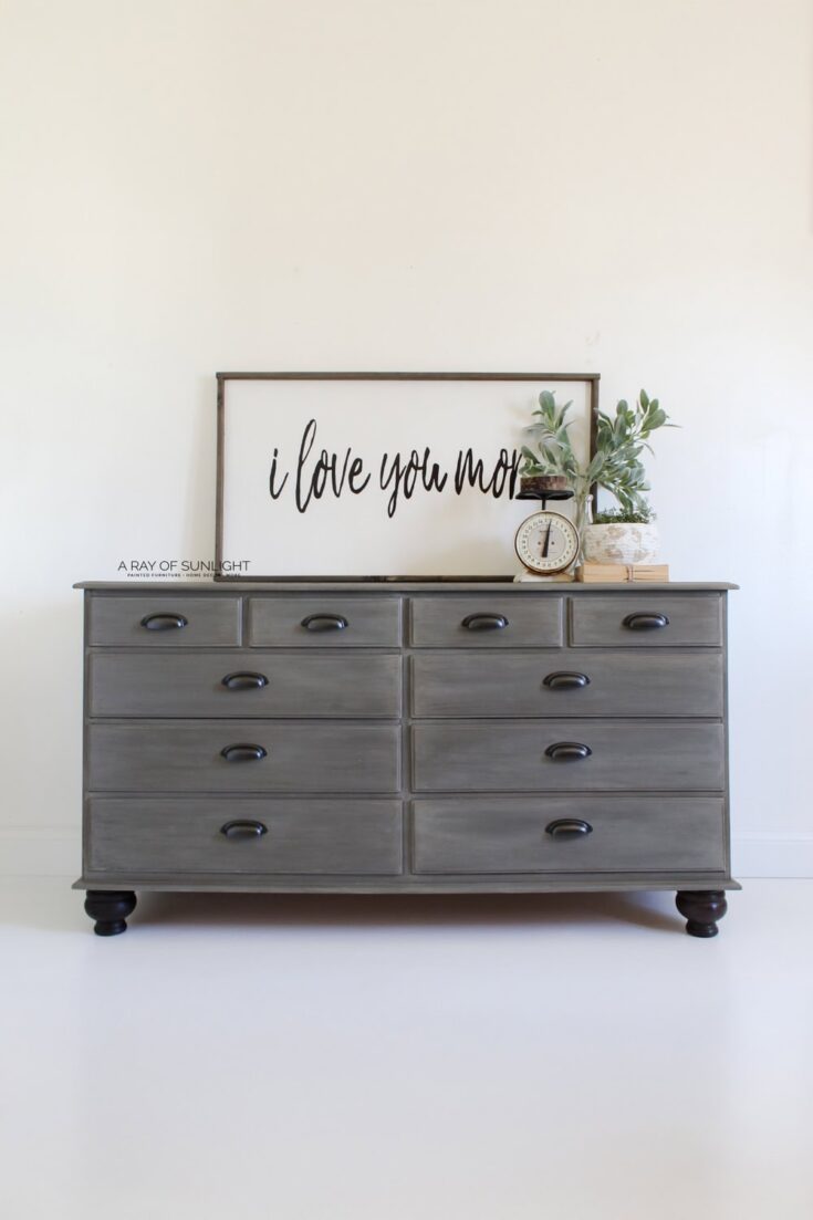 Gray dresser with six drawers, decor items, and a framed I love you more sign on top against a white wall.