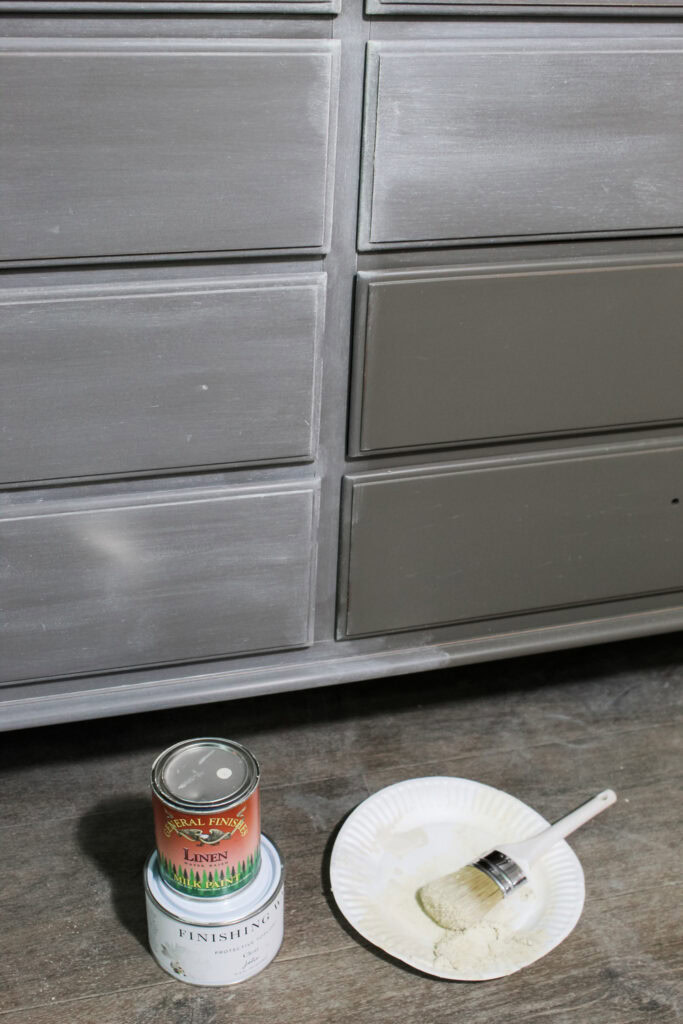 Dresser with a weathered paint finish, with paint and wax cans, and a brush on a paper plate on the floor.