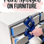 A person uses a blue paint sprayer on a dresser with drawers taped off for painting.