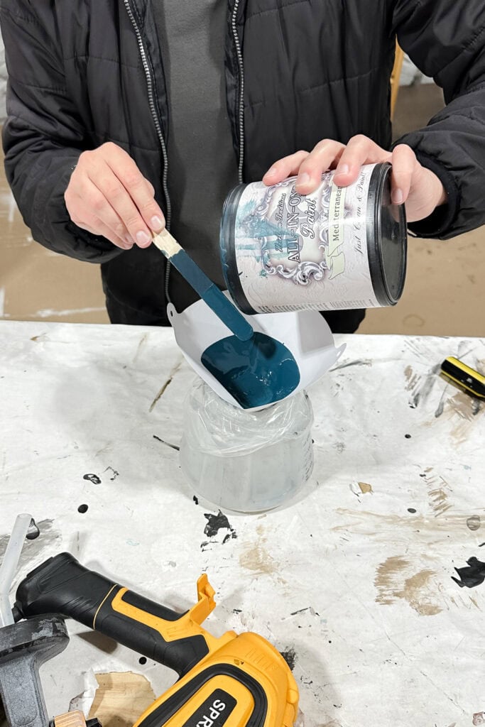 Person pouring dark blue paint from a can into sprayer container.