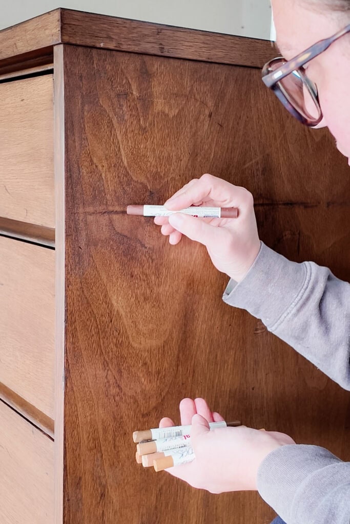 Person using a wood stain marker to cover scratches on wood, holding markers in other hand.
