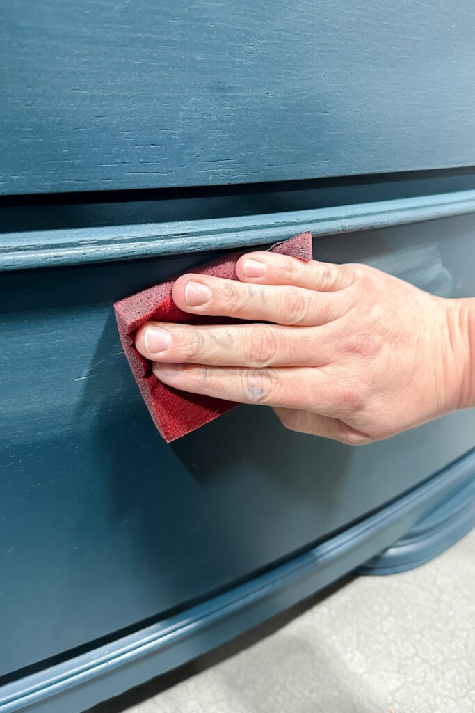 A hand uses a red foam sanding pad to smooth the surface of blue painted wood furniture.