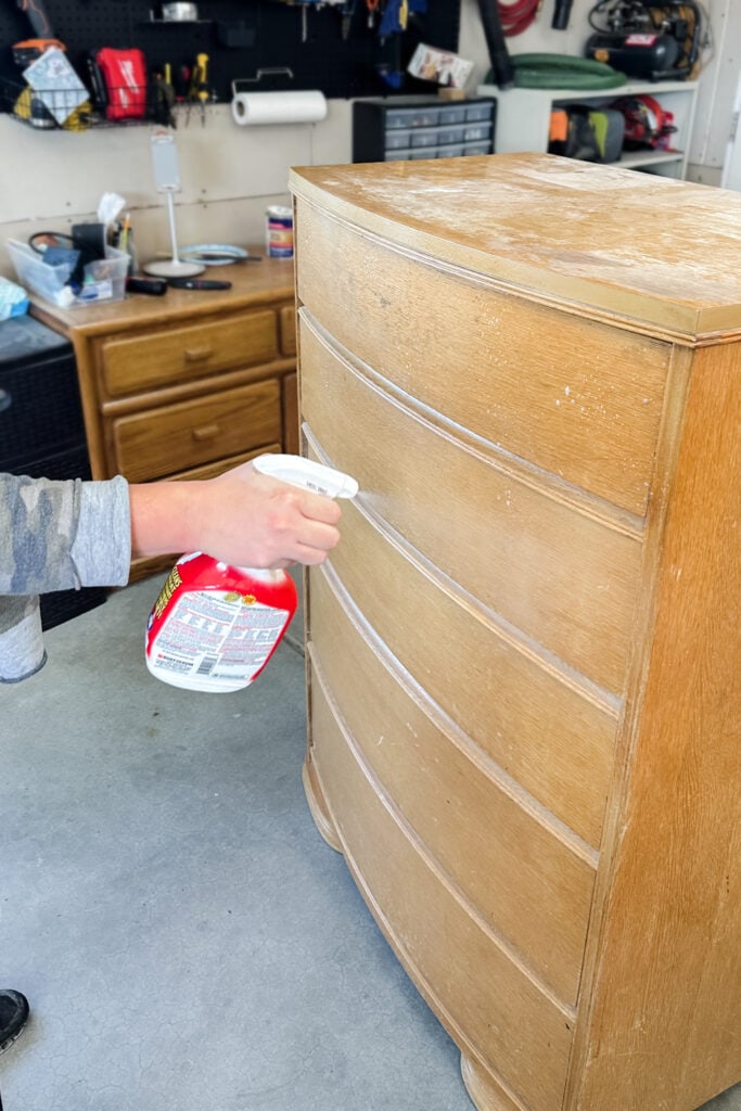 Person spraying cleaner onto a dusty wooden dresser in a garage
