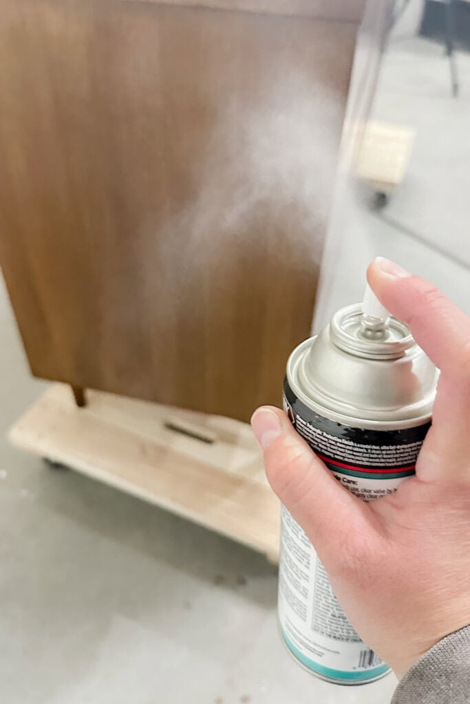 A hand sprays topcoat from a can onto a wooden dresser