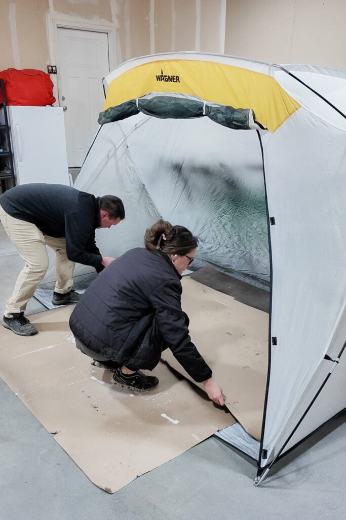 Two people set up cardboard sheets inside a portable spray tent in a garage.