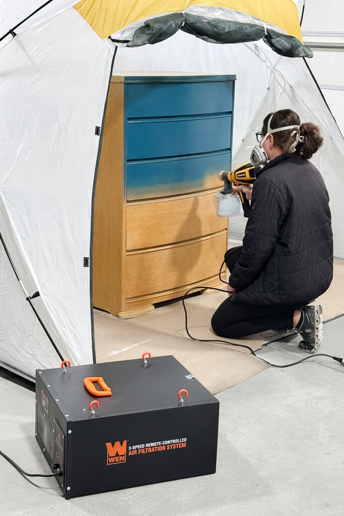 Person wearing a mask spray paints a dresser blue inside a tent with an air filtration system nearby.