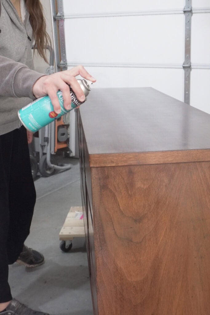 A person sprays topcoat on a wooden dresser in a garage workshop setting.