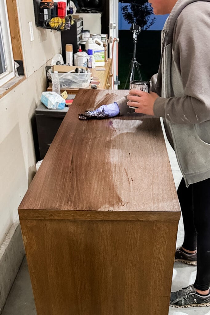 Person applying stain to a wooden dresser top in a garage