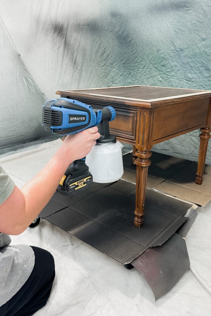 Person using a paint sprayer to refinish a wooden table, with cardboard protecting the floor.