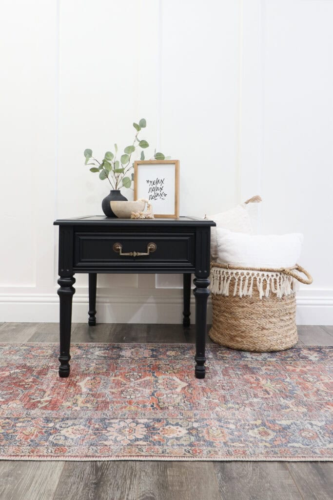 Black side table with decor, next to a woven basket with white pillows, on a patterned rug.
