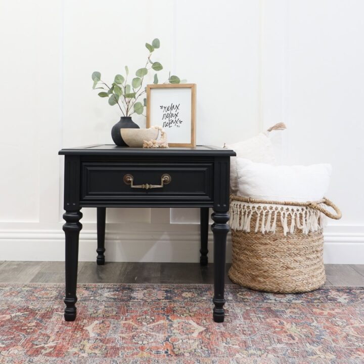 Black side table with decor, next to a woven basket with white pillows, on a patterned rug.
