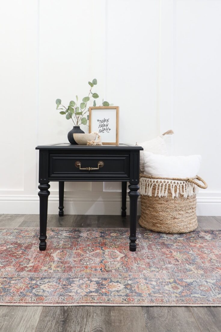 Black side table with decor, next to a woven basket with white pillows, on a patterned rug.