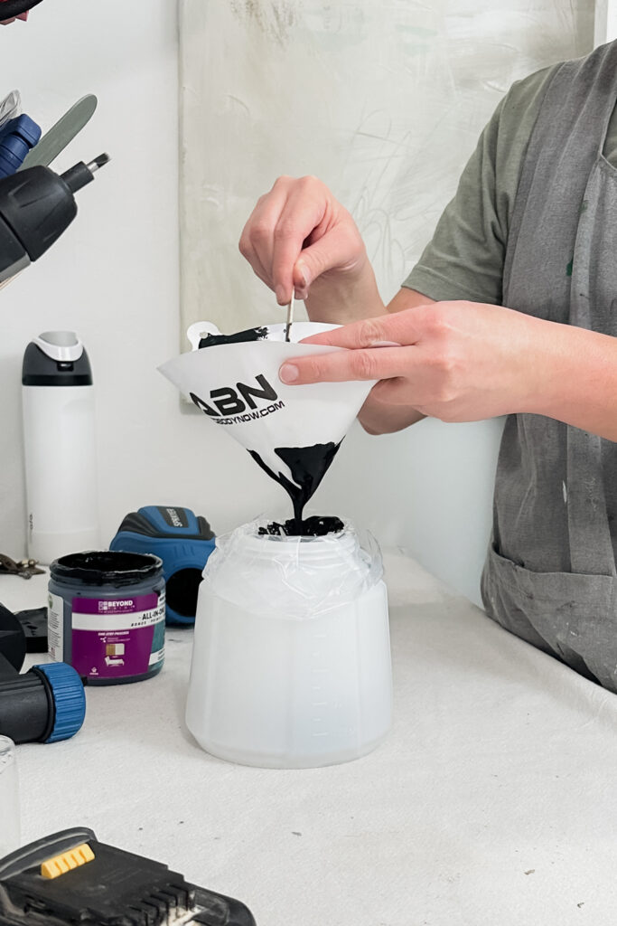 Person pouring black liquid through a funnel into a plastic container on a worktable.