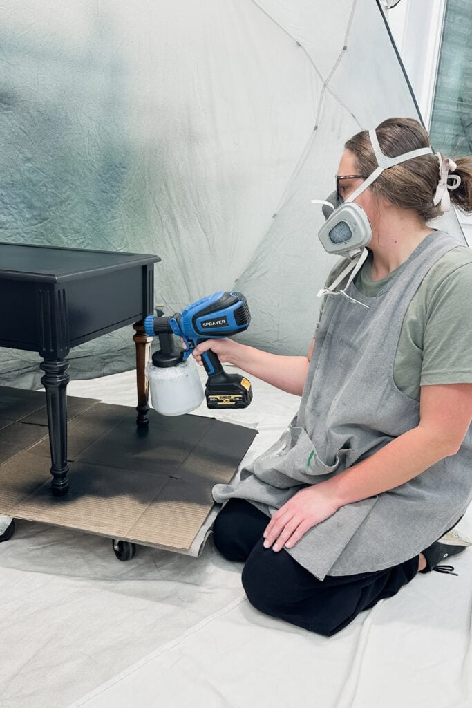 A woman wearing a mask uses a paint sprayer to paint a black side table indoors.