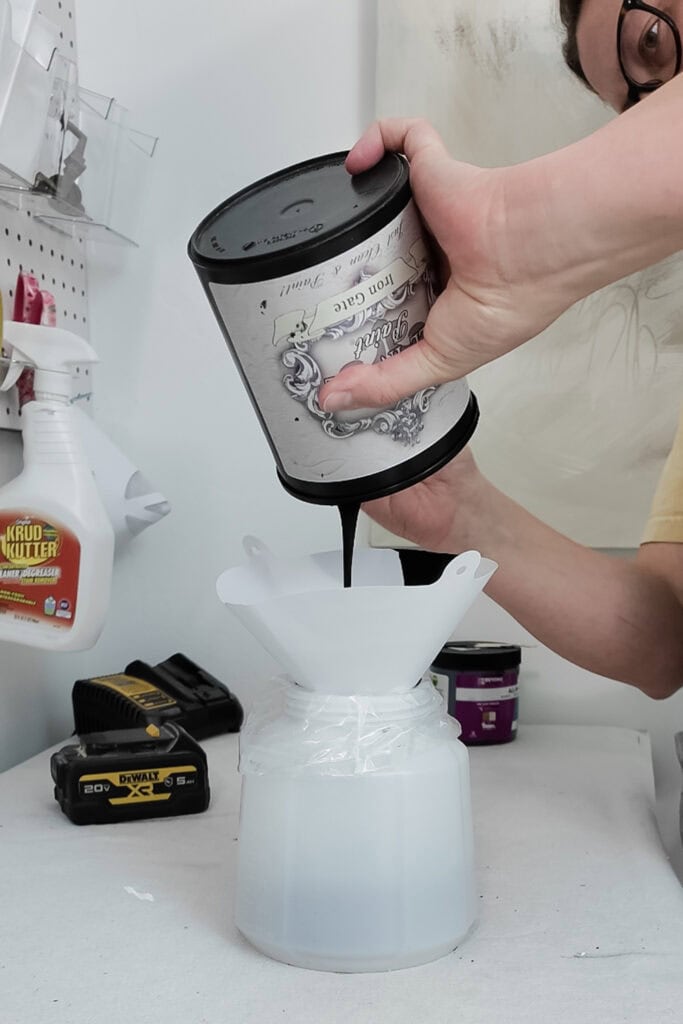 A person pours black paint from a can through a funnel into a large white container.
