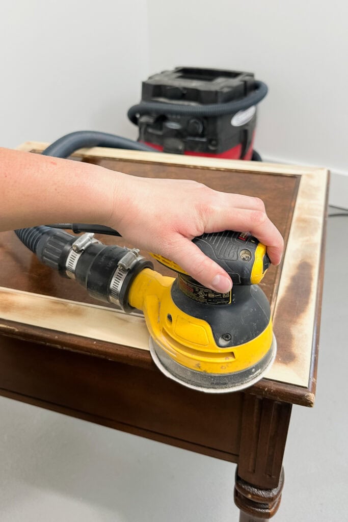 A hand uses a yellow electric sander on a wooden table with a vacuum in the background.