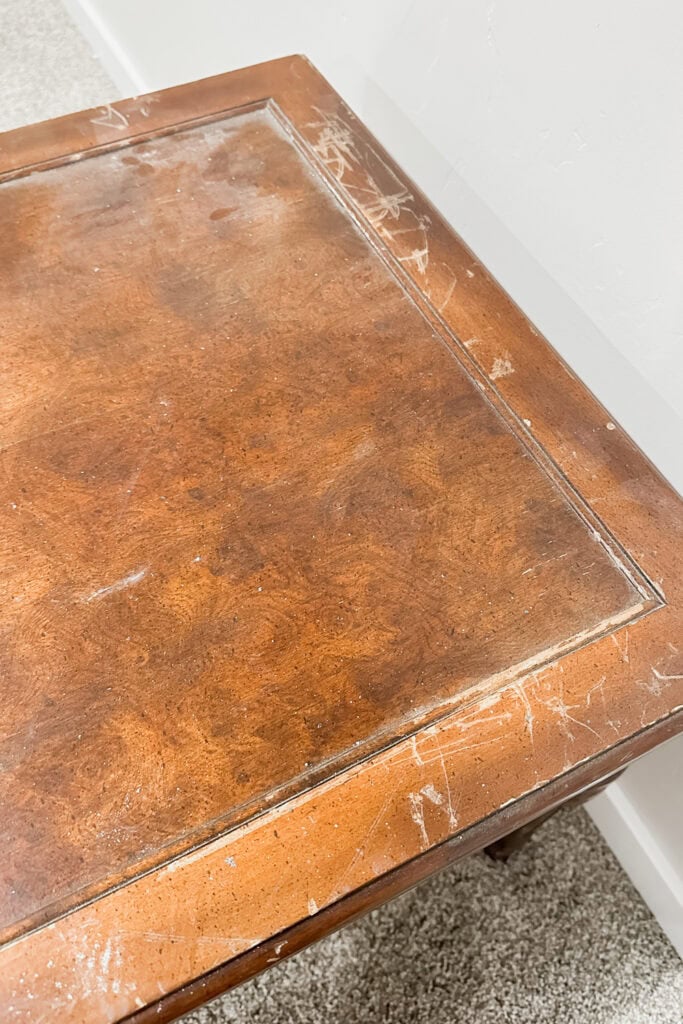 A wooden table with visible scratches and scuff marks on its surface, near a white wall and carpeted floor.